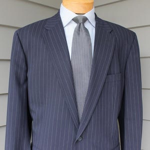 May include: A navy blue pinstripe suit jacket with a matching tie and white dress shirt. The suit is tailored with a notched lapel and a single-button closure. The tie is a solid gray color.
