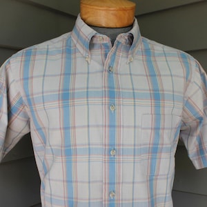 May include: A short-sleeved button-down shirt in a plaid pattern. The shirt features a white base with blue and red lines forming the plaid. It has a button-down collar and a chest pocket. The shirt is displayed on a wooden mannequin.
