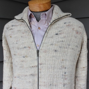 May include: A cream-colored zip-up sweater with a textured knit pattern. The sweater has flecks of color throughout and a high collar. A striped button-down shirt is visible underneath.