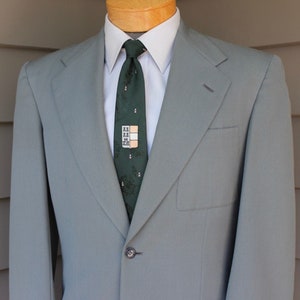 May include: A light gray blazer with a single button closure and a dark green patterned tie. The tie has a small, rectangular design with a light brown background.