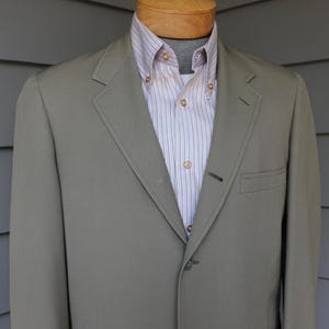 May include: A solid sage green blazer with notched lapels and a single-button closure, worn over a light-colored button-down shirt featuring thin vertical stripes in shades of blue and red, displayed on a mannequin torso.