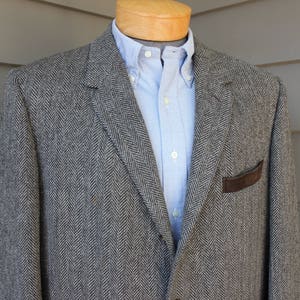 May include: A gray herringbone blazer with a brown leather patch on the left pocket. The blazer has a light blue button-down shirt underneath.