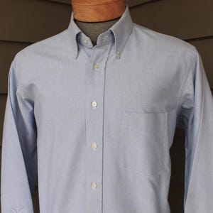 May include: Light blue button-down shirt with a button-down collar and a chest pocket. The long-sleeved shirt is displayed on a mannequin. The shirt has a classic, tailored design.
