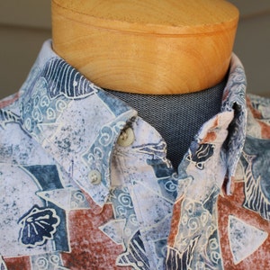 May include: A white button-down shirt with a blue, brown, and white abstract print. The shirt has a collar and a button-up closure.