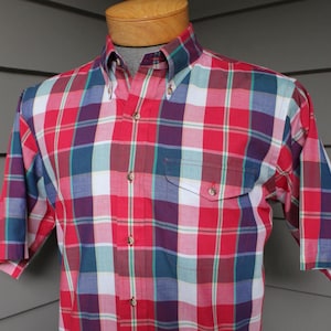 May include: A short-sleeved button-down shirt in a red, white, blue, and purple plaid pattern. The shirt has a button-down collar and a single chest pocket. The shirt is displayed on a mannequin.
