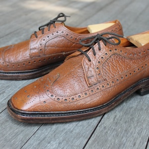 May include: A pair of brown leather brogue shoes with a wingtip design. The shoes have a lace-up closure and a textured leather upper.