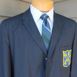 May include: A dark blue blazer with a single button closure and a patch pocket on the left side. The pocket features a yellow and blue embroidered crest with a lyre, a tree, and crossed swords.