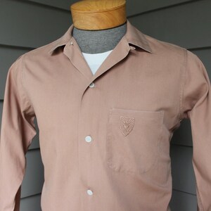 May include: A long-sleeved, button-down shirt in a muted rose color, featuring a chest pocket with an embroidered emblem. The shirt has a classic collar and is displayed on a mannequin.