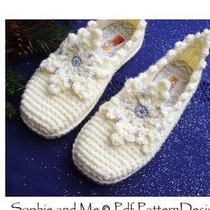 Può includere: Un paio di pantofole bianche all'uncinetto con motivi a fiocco di neve. Le pantofole hanno un accento di glitter argentato sui fiocchi di neve.