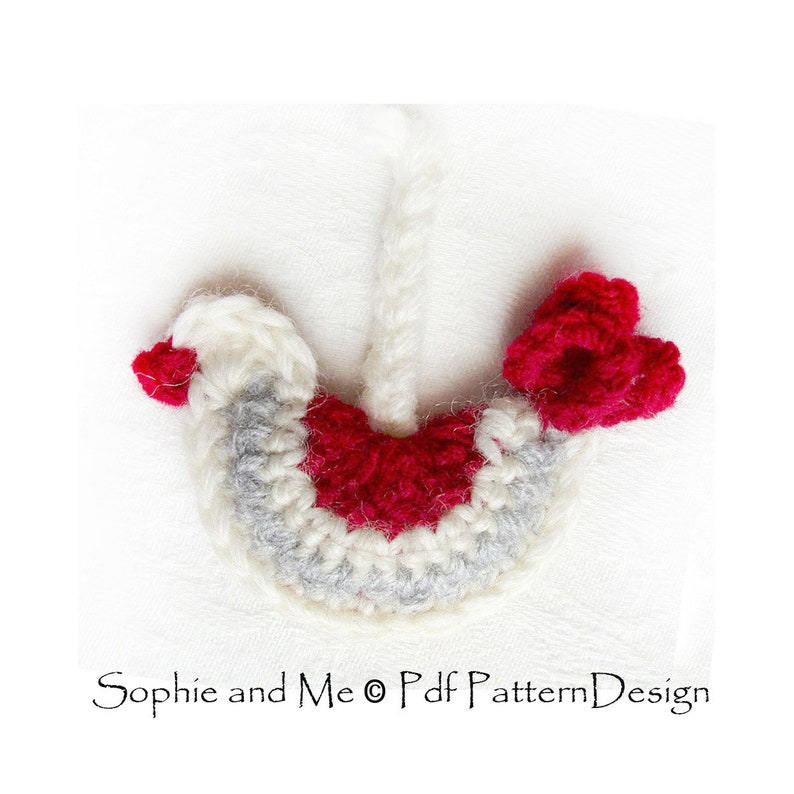 May include: A crocheted red and white bird ornament with a gray body and a red flower on the side. The ornament is hanging from a white string.