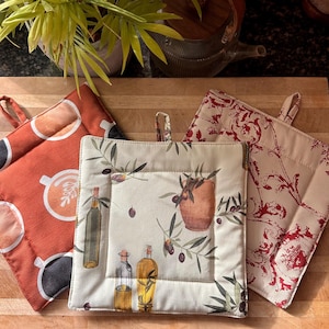 May include: Three square pot holders with loop handles. One is orange with a coffee cup design, another is cream with olive branches and oil bottles, and the third is cream with a red floral pattern. Kitchen textiles.