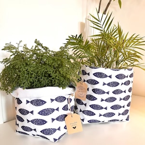 May include: Two white fabric storage baskets featuring a repeating dark blue fish pattern. Each basket has a folded top edge and a brown paper tag attached with twine. One tag reads "made with" followed by a heart symbol, and the other says "thank you."