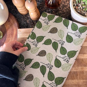 May include: A beige linen kitchen towel with a repeating pattern of green, brown, and off-white leaves. The towel has a handwritten script along the edges. The towel is on a wooden cutting board.