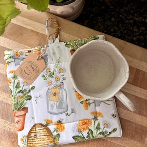 May include: A white fabric pot holder featuring a pattern of yellow dandelion flowers, green leaves, a glass jar, and a beehive, with a beige paper tag labelled Simplylinathreads thank you homemade with love, and a speckled white ceramic mug.