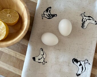 May include: A beige kitchen towel with black chicken illustrations and two white eggs. The towel is placed on a wooden cutting board next to a wooden bowl with lemon halves.