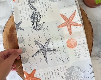 May include: A white kitchen towel with a seashell and starfish pattern. The design includes seahorses, starfish, and seashells in shades of gray and orange, with a script-like background. The towel is folded and resting on a wooden surface.