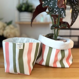 May include: Two fabric storage baskets featuring vertical stripes in muted olive green, soft coral, and white. The baskets have a white interior lining and a small rectangular tag on the front of the left basket that reads handmade with love.