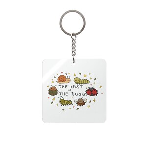 Pode incluir: Um porta-chaves branco com um anel e corrente prateados. O porta-chaves apresenta uma ilustração colorida de vários insetos de desenho animado, incluindo um caracol, uma lagarta, uma joaninha e outros, com o texto "THE LAST OF THE BUGS".