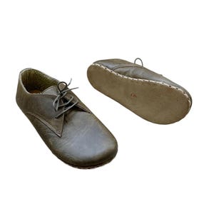 May include: A pair of olive green leather shoes with laces. One shoe is upright, and the other is lying on its side, showing the sole. The soles are stitched with white thread.