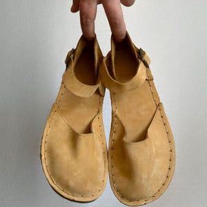 May include: A pair of light tan leather sandals with a strap and buckle closure. The sandals have a rounded toe and are held up by a hand. The stitching is visible around the edges of the sole.