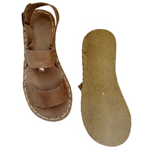 May include: Brown leather sandals with a strap around the ankle and a toe loop. The sandals have a stitched design around the edges and a buckle closure. The sole is a lighter brown color.