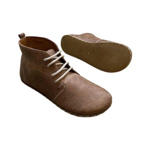Op de afbeelding: Bruine leren enkellaarsjes met witte veters. De laarzen hebben een ronde neus en een treklus aan de achterkant. De zool van één laars is zichtbaar en toont de stiksels.