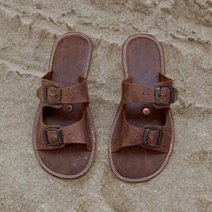 May include: A pair of brown leather sandals with two adjustable straps and metal buckles. The sandals have a stitched white sole and are displayed on a sandy surface. The footwear is designed for casual wear.