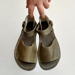 May include: A pair of olive green leather sandals with a strap and buckle closure. The sandals have a rounded toe and are held up by a hand against a white background. The edges are stitched with white thread.