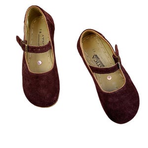 May include: A pair of burgundy suede Mary Jane style shoes. The shoes have a strap with a buckle closure and a rounded toe. The interior is a light tan color, and the sole is a natural color. The brand name "ATICA" is visible on the strap.