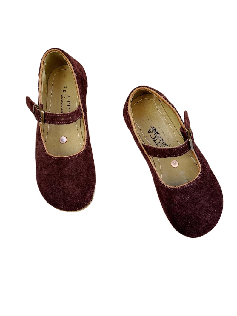 May include: A pair of burgundy suede Mary Jane style shoes. The shoes have a strap with a buckle closure and a rounded toe. The interior is a light tan color, and the sole is a natural color. The brand name "ATICA" is visible on the strap.