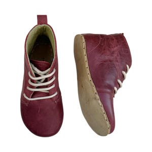 Könnte beinhalten: Zwei burgunderfarbene Leder-Ankle Boots mit cremefarbenen Schnürsenkeln. Die Stiefel haben eine runde Spitze und eine Lasche hinten. Die Sohlen sind hellbraun. Die Stiefel werden vor weißem Hintergrund gezeigt.