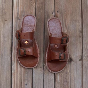 May include: A pair of brown leather sandals with two adjustable straps and metal buckles. The sandals have a stitched white sole and are displayed on a wooden surface. The footwear is designed for casual wear.