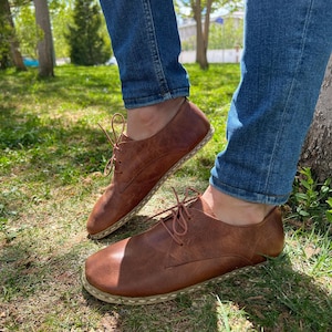 Op de afbeelding: Bruine leren schoenen met veters. De schoenen hebben een ronde neus en een gestikte zool. De afbeelding toont de schoenen die worden gedragen met een blauwe spijkerbroek op een grasachtig oppervlak.