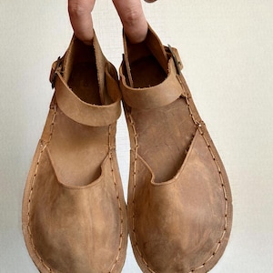 May include: A pair of brown leather sandals with a strap and buckle closure. The sandals have a rounded toe and are hand-stitched around the edges. The leather has a worn, textured appearance.