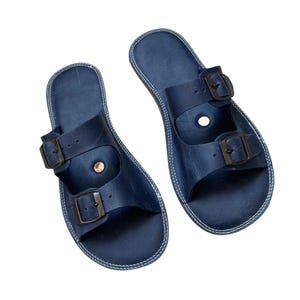 May include: A pair of navy blue leather sandals with two adjustable straps and metal buckles. The sandals have a contoured footbed and white stitching around the edges. Footwear.