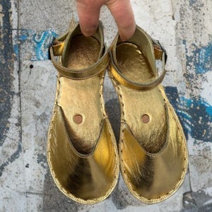 May include: A pair of gold-colored sandals with a metallic sheen. The sandals have a closed-toe design with a strap around the ankle and a buckle closure. The soles are stitched with a contrasting thread.