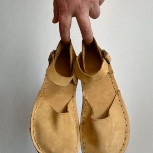 May include: A pair of light tan leather sandals with a strap and buckle closure. The sandals have a rounded toe and are held up by a hand. The stitching is visible around the edges of the sole.
