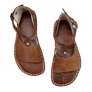 May include: A pair of brown leather sandals with ankle straps and buckle closures. The sandals have a stitched design around the edges and a small metal button detail. The leather has a textured appearance.