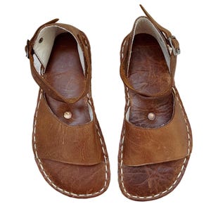 May include: A pair of brown leather sandals with white stitching. The sandals have an ankle strap with a buckle and a toe strap. The soles are a darker brown, and the sandals appear to be handmade.