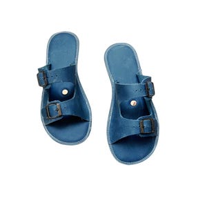 May include: A pair of blue leather sandals with two adjustable buckle straps. The sandals have a contoured footbed and a stitched edge. The buckles are a dark metal color. The sandals are shown on a white background.