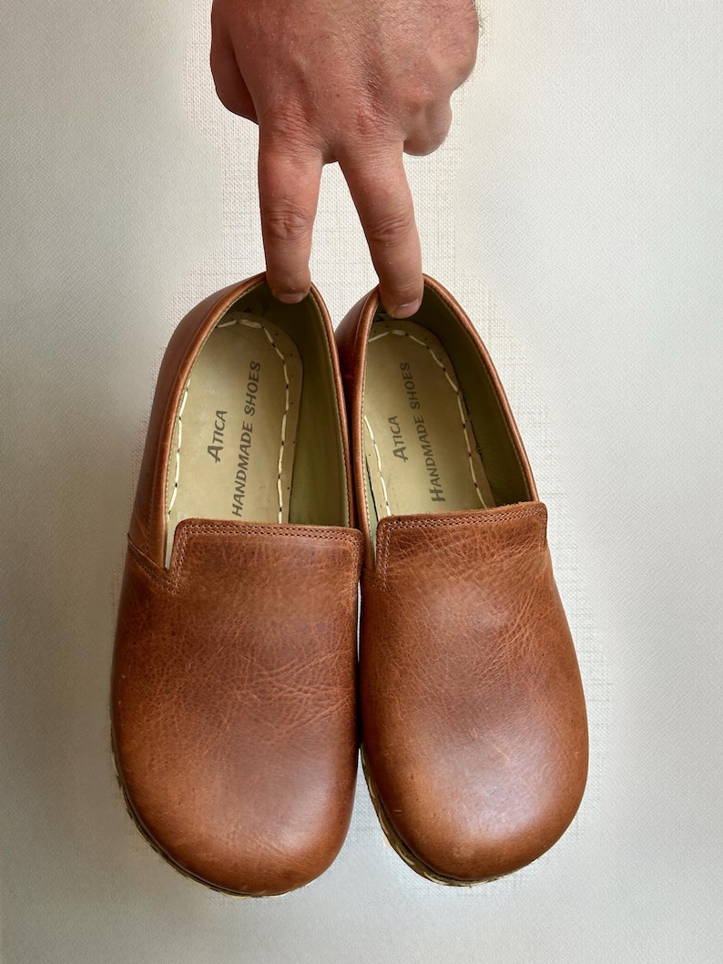 May include: A pair of brown leather slip-on shoes. The shoes have a rounded toe and a textured surface. The brand name "Atica Handmade Shoes" is printed on the inner lining. The shoes are held up by a person's fingers.