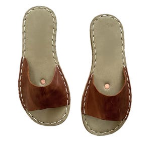 May include: A pair of handmade leather sandals. The sandals have a light green sole with a brown leather strap secured with a copper rivet. The edges are stitched with white thread.