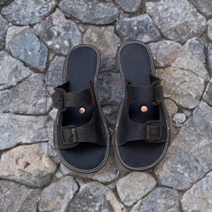 May include: A pair of black leather sandals with two adjustable buckle straps. The sandals have white stitching around the edges and are set against a background of gray stones.