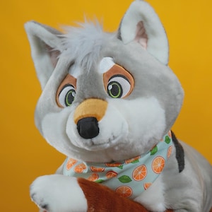 May include: A plush wolf toy with gray fur and white accents, wearing a light blue bandana patterned with orange slices. The wolf is posed on a round wooden table with a black metal base, set against a yellow background. The toy has green eyes and a black nose.