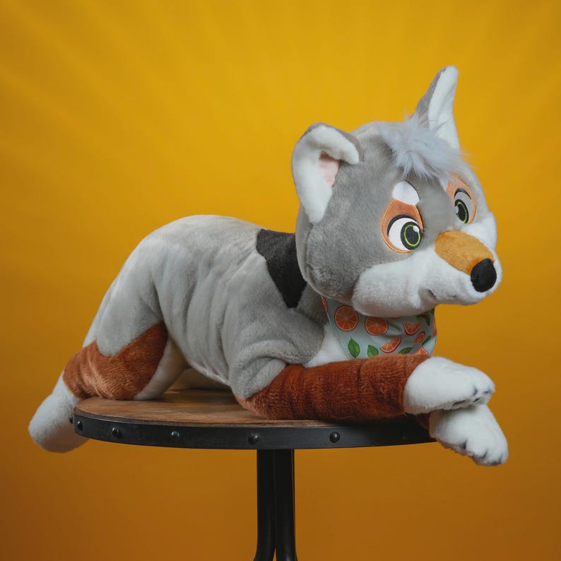 Peut inclure: Une peluche en forme de loup dans les tons de gris, marron et blanc, avec une tache noire sur le dos. Le loup porte une &eacute;charpe vert clair avec des tranches d'orange. Il est pos&eacute; sur une petite table ronde marron avec un fond jaune.