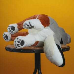 Peut inclure: Une peluche en forme de chien dans une pose ludique, avec une fourrure marron, blanche et grise. Les pattes du jouet sont visibles, avec des coussinets noirs. Le chien est pos&eacute; sur une petite table ronde en bois avec une base en m&eacute;tal noir, sur fond jaune.
