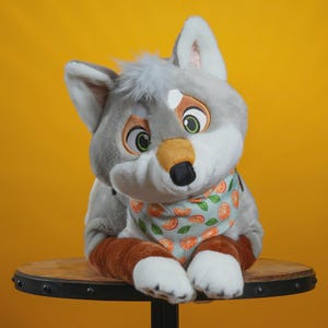Peut inclure: Une peluche en forme de loup avec une fourrure grise, des accents blancs et un bandana bleu clair orn&eacute; de tranches d'orange. Le loup a de grands yeux expressifs et est assis sur une surface en bois sur fond jaune.