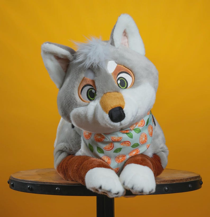Peut inclure: Une peluche en forme de loup avec une fourrure grise, des accents blancs et un bandana bleu clair orn&eacute; de tranches d'orange. Le loup a de grands yeux expressifs et est assis sur une surface en bois sur fond jaune.