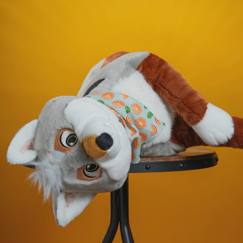 Peut inclure: Une t&ecirc;te d'animal en peluche grise et blanche, avec des oreilles marron et un nez noir. Elle pr&eacute;sente de grands yeux et une &eacute;charpe bleu clair orn&eacute;e de tranches d'orange. La t&ecirc;te repose sur un tabouret noir sur fond jaune.