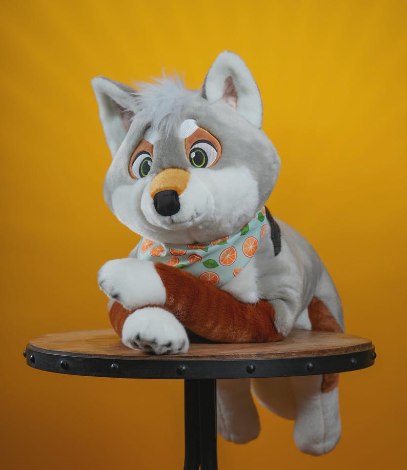 May include: A plush wolf toy with gray fur and white accents, wearing a light blue bandana patterned with orange slices. The wolf is posed on a round wooden table with a black metal base, set against a yellow background. The toy has green eyes and a black nose.